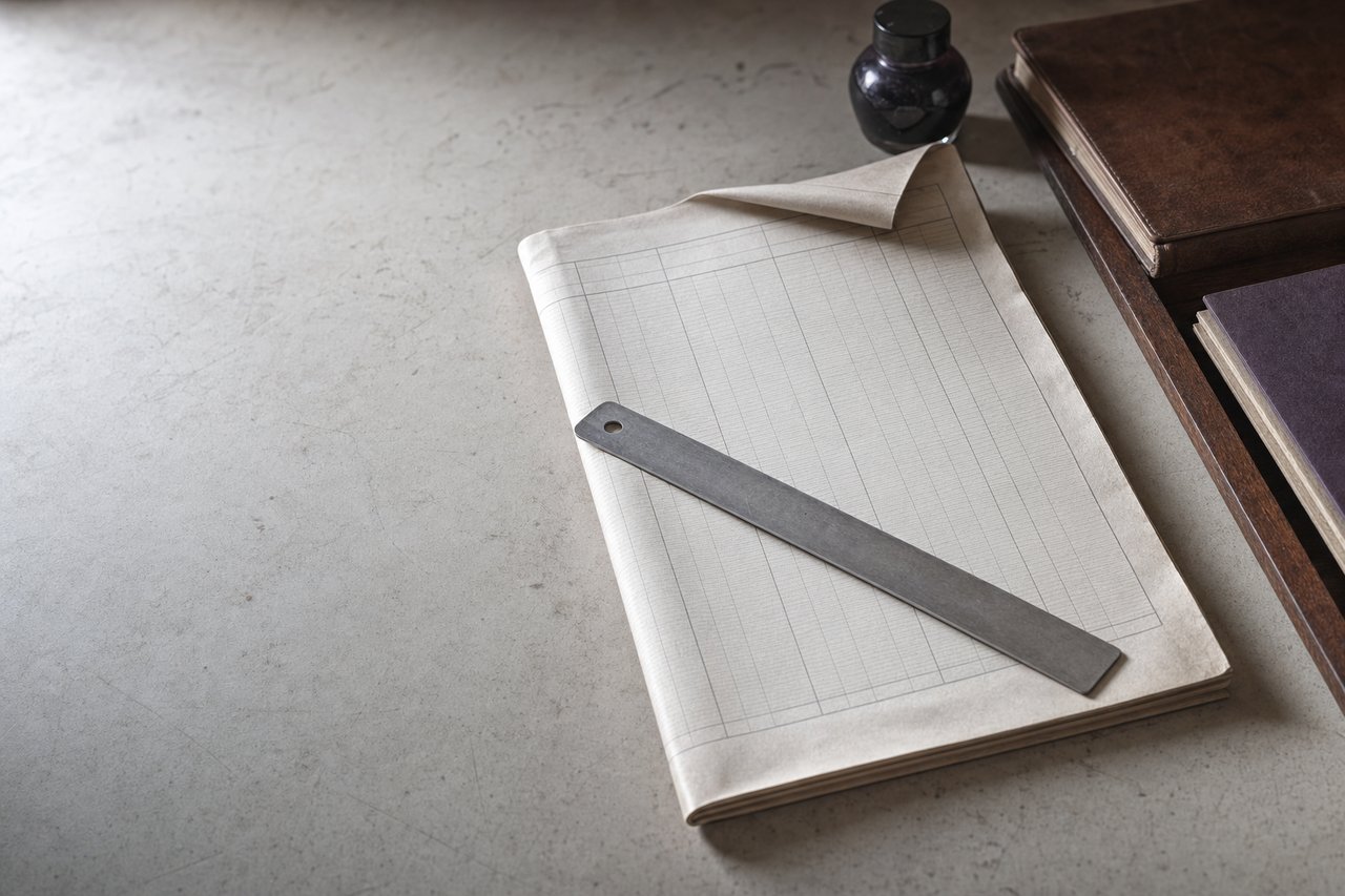 Pliego doblado de papel con retícula estadística en blanco sobre una mesa clara, con una regla metálica atravesada y una esquina levantada.